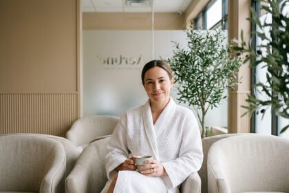 Femme détendue en robe blanche dans une salle d'attente de clinique esthétique moderne, tenant une tasse de tisane dans un cadre apaisant